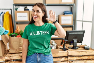 Bağışta gönüllü tişört giyen genç esmer kadın selam veriyor mutlu, güler yüzlü, dostça bir jest yapıyor. 