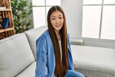 Genç Çinli kız gülümsüyor. Evdeki kanepede mutlu mesut oturuyor..