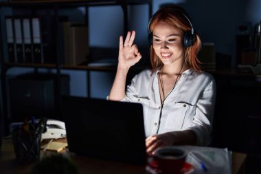 Geceleri ofiste çalışan beyaz bir kadın olumlu gülümsüyor ve el ve parmaklarıyla 