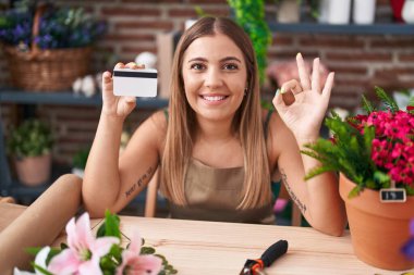 Young blonde woman working at florist shop holding credit card doing ok sign with fingers, smiling friendly gesturing excellent symbol 