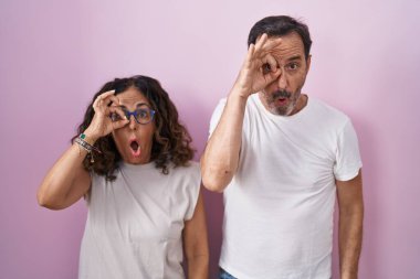 Orta yaşlı İspanyol çift pembe arka planda birlikte hareket ediyorlar. Şaşırmış bir yüz ve parmaklarından bakan gözler. İnançsız ifade. 