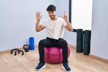 Sakallı İspanyol bir adam yoga odasında pilatese oturmuş yedinci parmaklarıyla işaret ederken kendinden emin ve mutlu bir şekilde gülümsüyor.. 