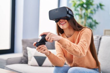 Evde sanal gerçeklik gözlüğü ve joystick kullanan güzel İspanyol bir kadın oyun oynuyor.