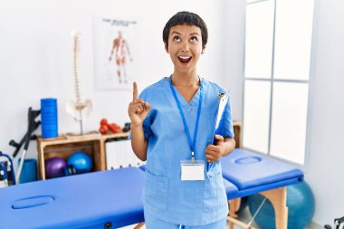 Kısa saçlı, İspanyol bir kadın ağrı kesici kliniğinde çalışıyor. Şaşırmış ve şaşırmış bir şekilde yukarı bakıyor, parmaklarıyla işaret ediyor ve kollarını kaldırıyordu.. 