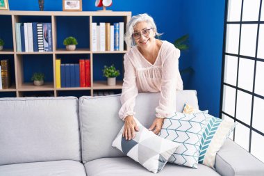 Orta yaşlı, gri saçlı, gülümseyen, kendine güvenen, evdeki kanepeyi organize eden kadın.