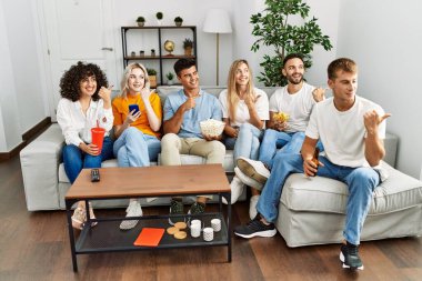 Bir grup insan evdeki kanepede oturmuş gülümseyen bir şekilde başparmağını kaldırıyor. 