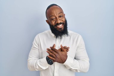 Mavi arka planda duran Afrikalı Amerikalı adam elleri göğsünde, gözleri kapalı ve yüzünde minnet dolu bir jest ile gülümsüyor. sağlık kavramı. 