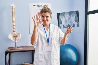 Elinde leğen kemiği radyografisi olan genç bir kadın parmaklarıyla imza atıyor, gülümseyerek mükemmel bir sembol oluşturuyor. 