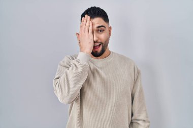 Genç yakışıklı adam izole edilmiş arka planda duruyor. Bir gözünü eliyle kapatıyor. Yüzünde kendine güvenen bir gülümseme ve şaşırtıcı bir duygu var.. 