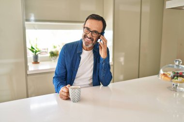 Orta yaşlı bir adam akıllı telefondan konuşuyor. Evde kahve içiyor.