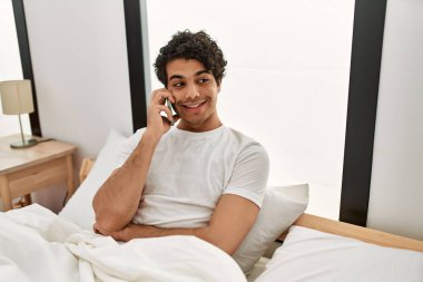 Genç İspanyol adam akıllı telefonda konuşuyor. Yatak odasında yatakta oturuyor..