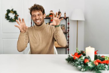 Masada sakallı genç yakışıklı bir adam Noel süslemelerinin yanında oturmuş komik bir şekilde gülüyor kedi gibi pençe hareketi yapıyor, agresif ve seksi bir ifade. 