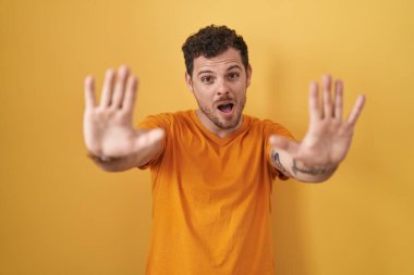 İspanyol asıllı genç adam sarı arka planda duruyor. Avuçlarıyla jestleri durduruyor. Öfkeli ve hayal kırıklığı dolu bir ifade. 