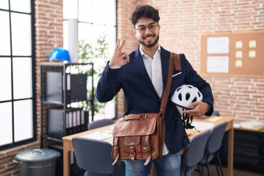 Ofiste sakallı, bisiklet kasklı İspanyol bir adam parmaklarıyla imza atıyor, gülümseyerek mükemmel bir sembol oluşturuyor. 