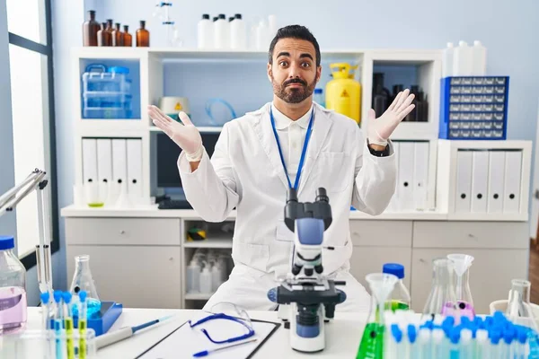 Young hispanic man with beard working at scientist laboratory clueless and confused expression with arms and hands raised. doubt concept.