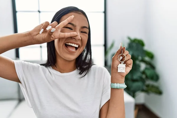 Young hispanic woman holding keys of new home doing peace symbol with fingers over face, smiling cheerful showing victory