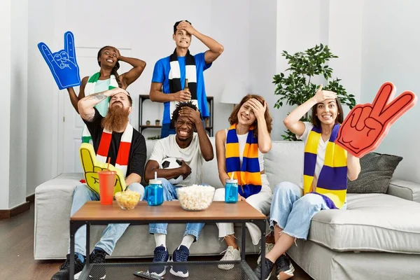 Group of friends supporting football team watching tv al home sitting on the sofa stressed and frustrated with hand on head, surprised and angry face