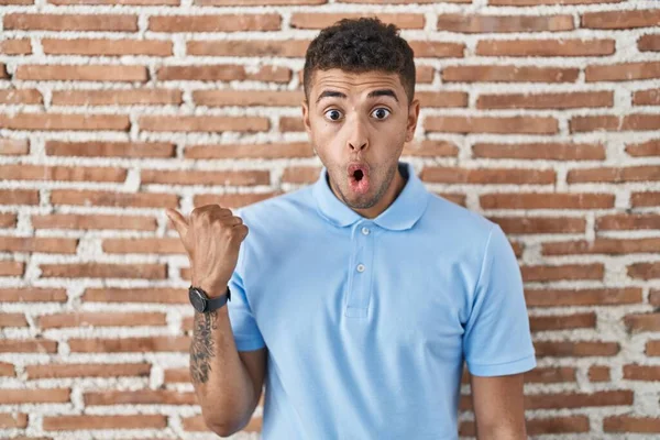 ブラジルの若い男がレンガの壁の上に立って驚いて手の指で側に指して 口を開けて驚いた表情 — ストック写真