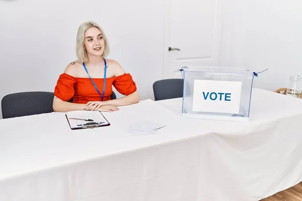 Young caucasian woman at political election sitting by ballot looking away to side with smile on face, natural expression. laughing confident.