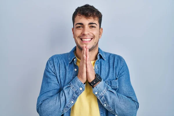 若いヒスパニック系の男が青の背景に立って手で一緒に笑みを浮かべて許しを求めて祈る自信を持って — ストック写真
