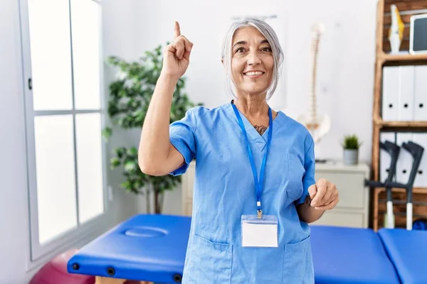 Medelålders Gråhårig Kvinna Bär Sjukgymnast Uniform Läkarmottagningen Pekar Finger Upp — Stockfoto