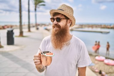 Genç, kızıl saçlı, gülümseyen, kumsalda kokteyl içen bir turist..