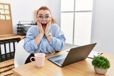 Ofiste çalışan genç kızıl saçlı kadın bilgisayar laptopu kullanıyor. Korkmuş, şok olmuş, şaşırmış ve hayrete düşmüş. 