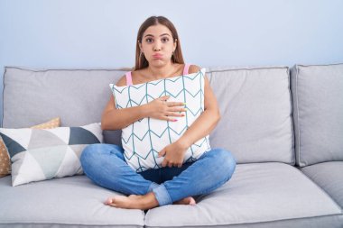 Young blonde woman hugging pillow sitting on the sofa puffing cheeks with funny face. mouth inflated with air, catching air. 