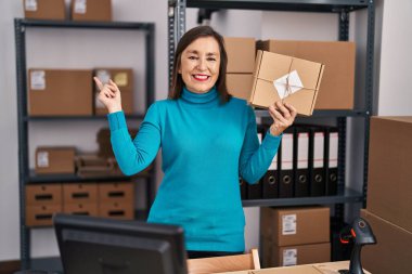 Orta yaşlı İspanyol bir kadın, küçük bir iş yerinde çalışıyor. Elinde bir koli var, gülümsüyor, mutlu bir şekilde elini ve parmağını yana uzatıyor. 