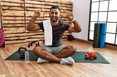 Spor salonundaki antrenman minderinde oturan genç Hintli adam şaşırmış, şaşırmış ve parmaklarıyla işaret ederek kollarını kaldırdı.. 
