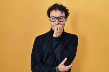 Sarı arka planda dikilen İspanyol adam gergin ve gergin görünüyor. Ağzı tırnak yiyen eller. anksiyete. 