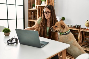 Genç İspanyol kadın evde köpekle masada dizüstü bilgisayar kullanıyor.