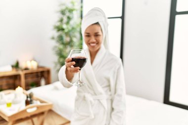 Bornoz giyen genç Latin kadın güzellik merkezinde şarapla kadeh kaldırıyor.