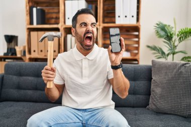 Sakallı, çekiçli ve akıllı telefonlu genç İspanyol adam kırık ekranı kızgın ve öfkeli bir şekilde bağırırken gösteriyor. Öfkeyle bağırıyor. öfke ve agresif konsept. 