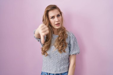 Pembe arka planda duran güzel sarışın kadın mutsuz ve kızgın görünüyor. Reddedilmiş ve olumsuz bir hareket yapıyor. kötü ifade. 