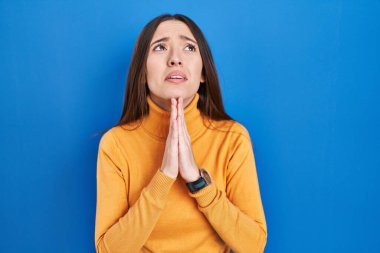 Genç esmer kadın mavi arka planda duruyor. Elleriyle yalvarıyor ve dua ediyor. Yüzünde umut ifadesi var. Yalvarıyor. 