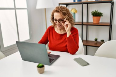 Orta yaşlı, beyaz bir kadın evdeki masada dizüstü bilgisayar kullanıyor..