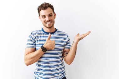 Yakışıklı genç adam izole edilmiş arka planda duruyor avuç içini gösteriyor ve baş parmağıyla iyi bir hareket yapıyor, mutlu ve neşeli gülümsüyor. 
