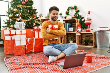 Genç Arap adam evdeki Noel ağacının yanında dizüstü bilgisayar kullanıyor..