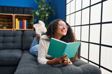 Evdeki kanepede kitap okuyan genç Latin kadın.