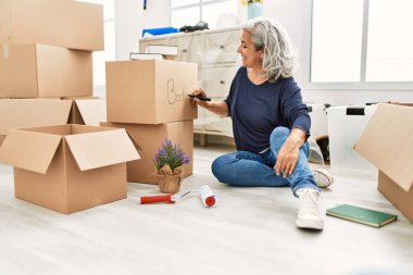 Middle age grey-haired woman writing books word on cardboard box at new home.