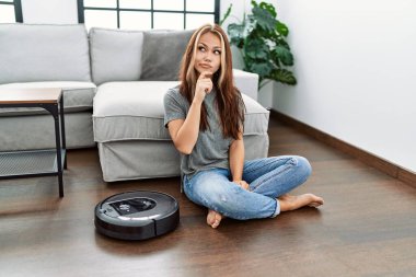 Evde vakumlu robot gibi oturan beyaz bir kadın ciddi bir yüzle eli çenesinde soru soran, kafa karıştırıcı bir fikir hakkında düşünceli. 