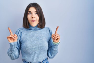 Mavi arka planda duran genç İspanyol kadın şaşırmış ve şaşırmış bir şekilde yukarı bakıyor ve parmaklarıyla işaret ediyor ve kollarını kaldırıyor.. 