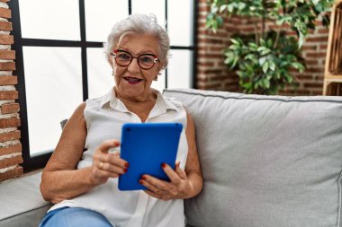 Yaşlı gri saçlı kadın evdeki kanepede oturuyor ve dokunmatik ped kullanıyor.