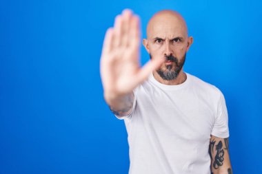 Mavi arka planda duran İspanyol dövmeli adam avucuyla şarkı söylemeyi kesiyor. Yüzünde olumsuz ve ciddi bir jest olan uyarı ifadesi. 