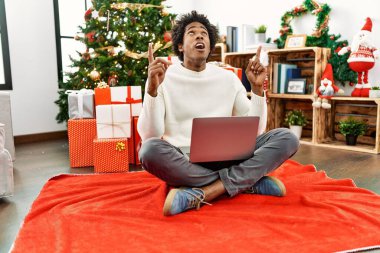 Genç Afro-Amerikan adam dizüstü bilgisayar kullanıyor. Yılbaşı ağacının yanında oturuyor. Şaşırmış ve şaşırmış.. 