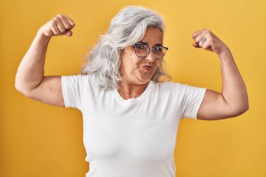 Sarı arka planda duran gri saçlı orta yaşlı kadın gururla gülümseyen kol kaslarını gösteriyor. fitness konsepti. 