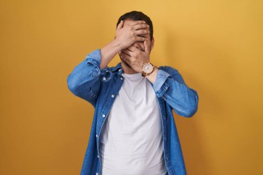 Sarı arka planda duran İspanyol adam gözleri ve ağzı elleriyle kapatıyor, şaşırmış ve şok olmuş. duygu gizleme 