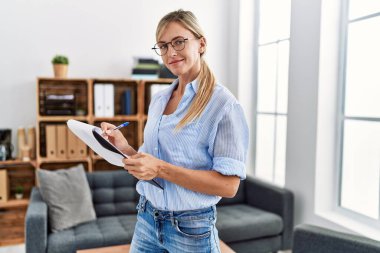 Young blonde woman psychologist writing on clipboard standing at clinic