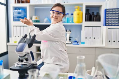 Down sendromlu İspanyol kız, bilim adamı laboratuarında çalışıyor, avuç içi açık, fotokopi alanını gösteren, gülümseyerek mutlu bir gülümseme sunan, 